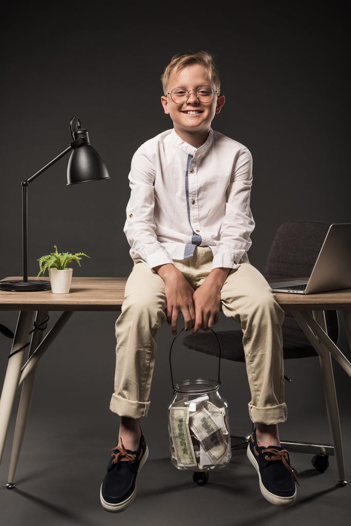 happy little boy in eyeglasses with jar full of dollar banknotes sitting on table with lamp, plant and laptop on grey background  - Photo, Image