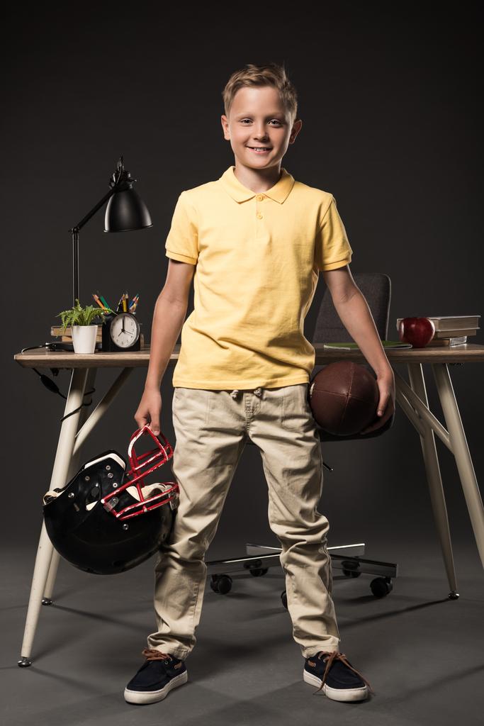 愛らしい少年ボールとアメリカン フットボールのヘルメットを押し書籍、植物、ランプ、色鉛筆、アップル、時計、灰色の背景の教科書でテーブルの近くに立っています。  - 写真・画像