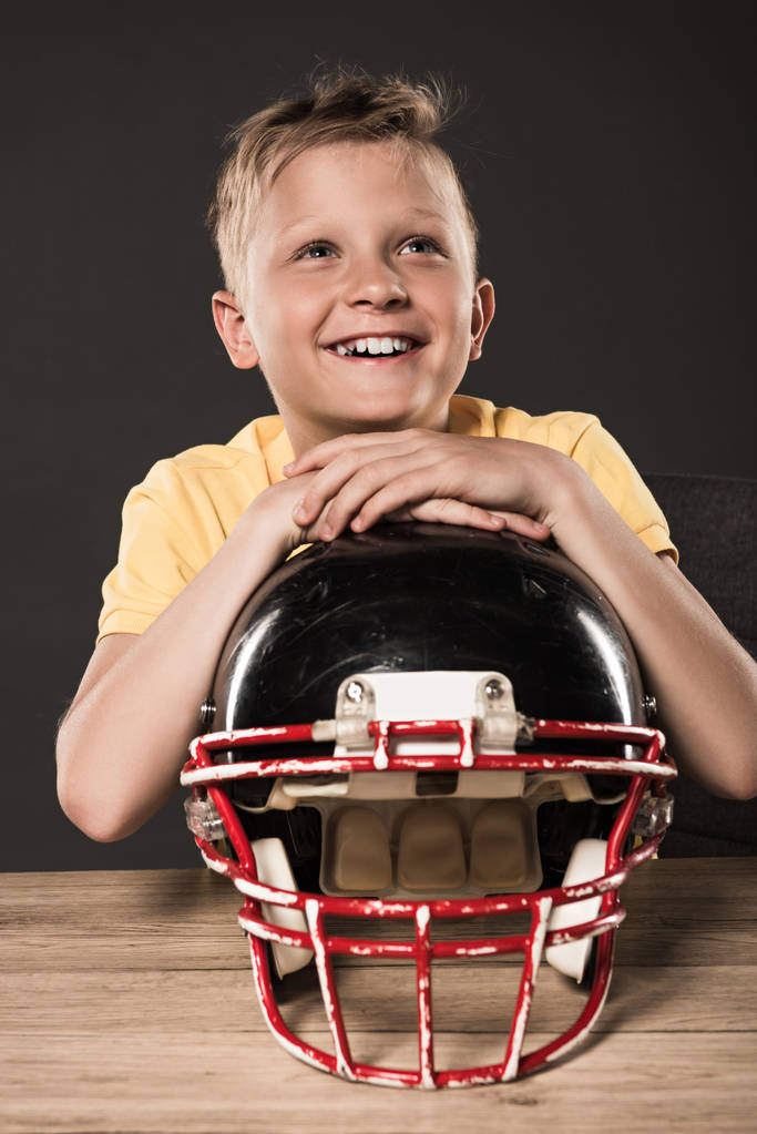 zasněný šťastný chlapeček sedí s helmu amerického fotbalu u stolu na šedém pozadí  - Fotografie, Obrázek