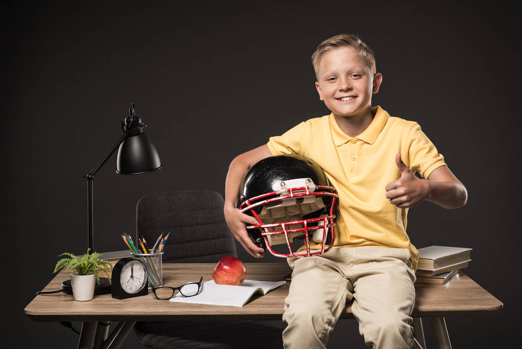 schoolboy holding american football helmet, doing thumb up gesture and sitting on table with books, eyeglasses, plant, lamp, colour pencils, apple, clock and textbook on grey background  - Photo, Image