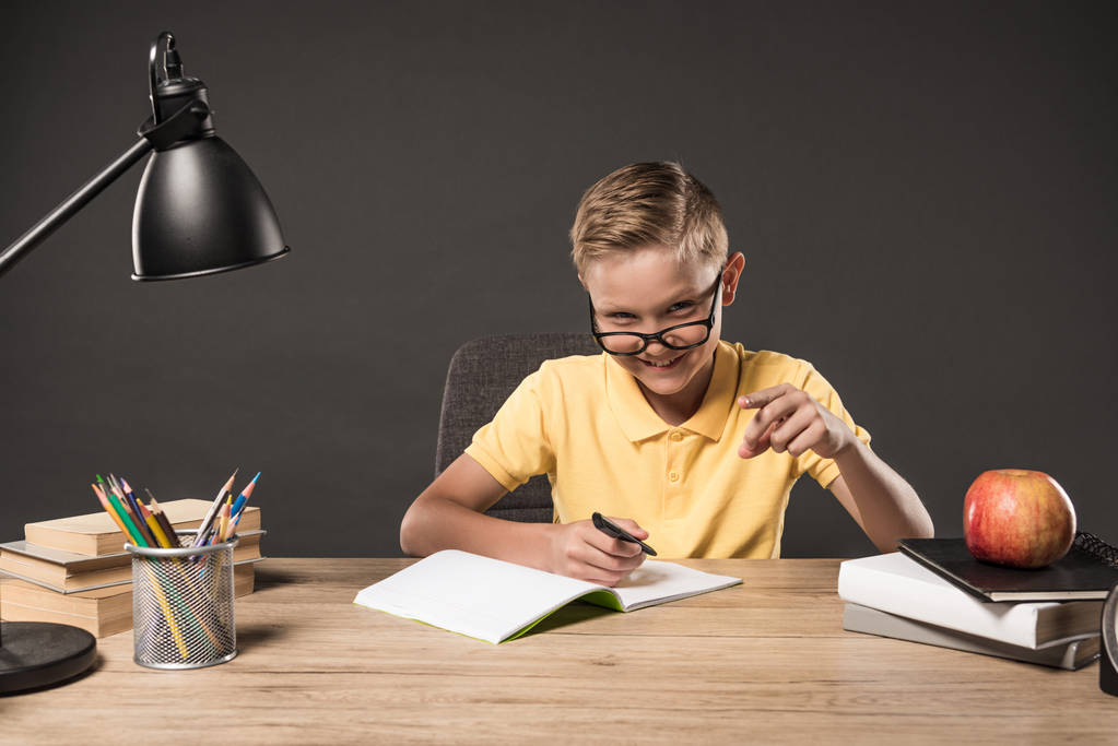 šťastný kluk v brýlové ukázal prstem a dělat domácí úkoly u stolu s lampou, knih, barevné tužky a učebnice na šedém pozadí  - Fotografie, Obrázek