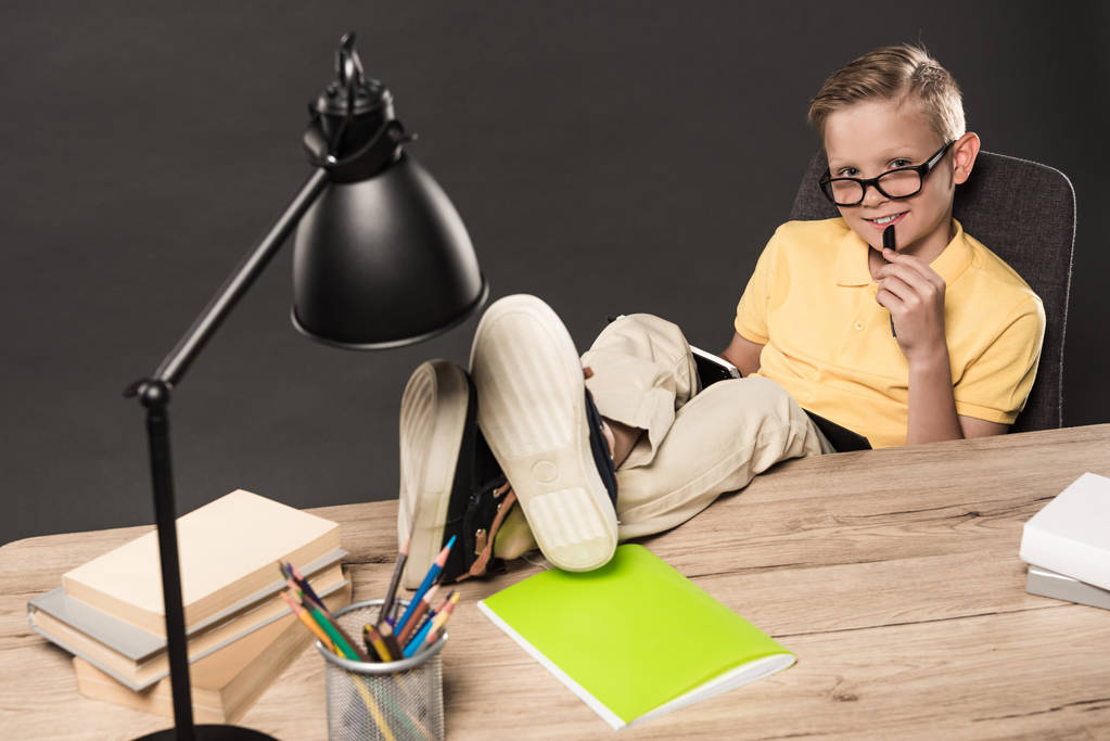 hymyilevä koulupoika silmälasissa tekemässä läksyjä jalat pöydällä lamppu, kirjat, värikynät ja oppikirja harmaalla taustalla
  - Valokuva, kuva