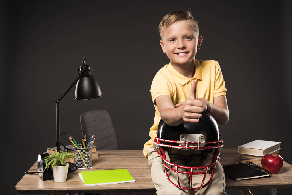 happy schoolboy holding american football helmet, doing thumb up gesture and sitting on table with books, plant, lamp, colour pencils, apple, clock and textbook on grey background  - Photo, Image