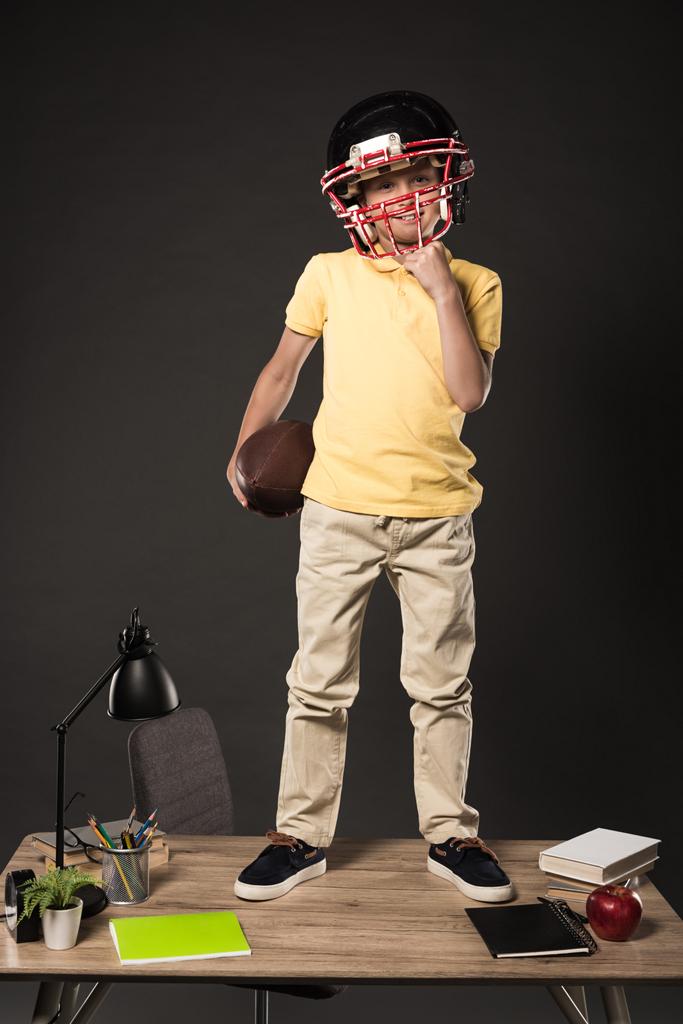 schoolboy in american football helmet holding ball and standing on table with books, plant, lamp, colour pencils, apple, clock and textbook on grey background  - Photo, Image