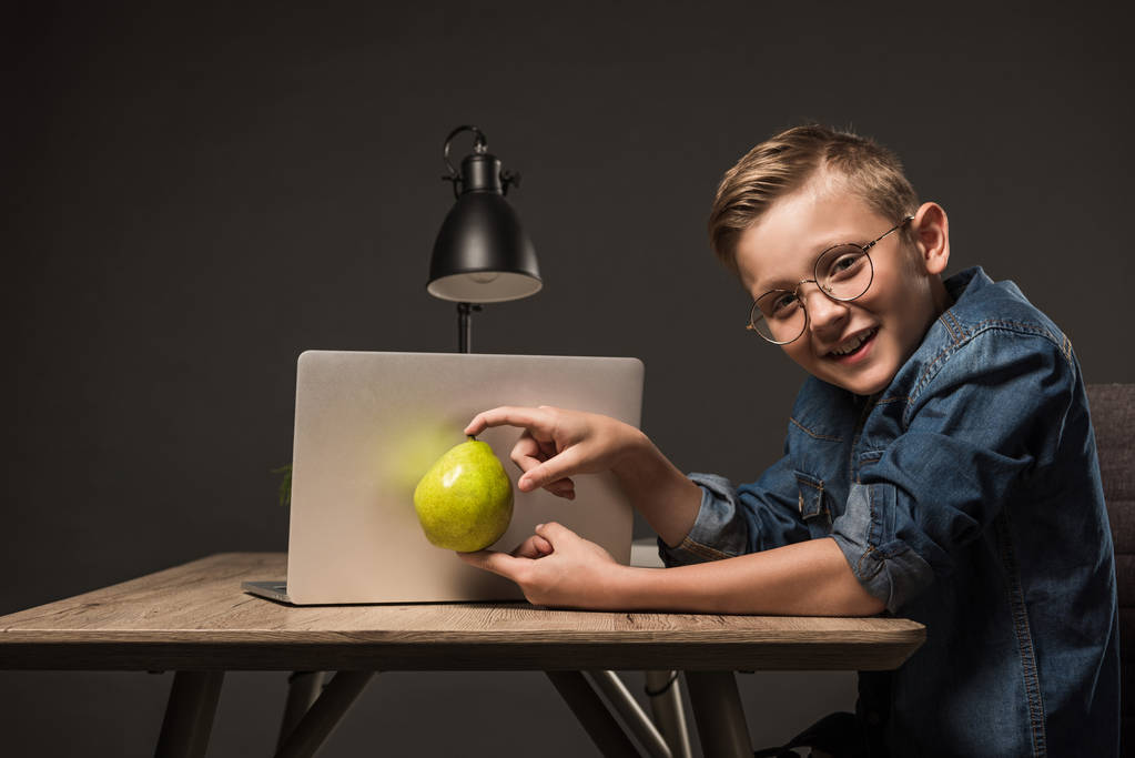 Lächelnder kleiner Junge in Brille mit Birne vor Laptop am Tisch mit Laptop auf grauem Hintergrund  - Foto, Bild