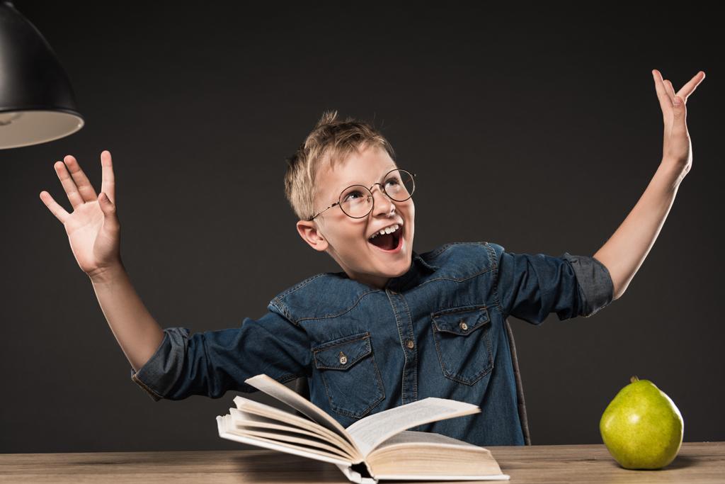 ενθουσιασμένος αγόρι σχολείο σε γυαλιά χειρονομώ από χέρια ενώ ανάγνωση βιβλίων στο τραπέζι με λάμπα και αχλάδι σε γκρίζο φόντο  - Φωτογραφία, εικόνα