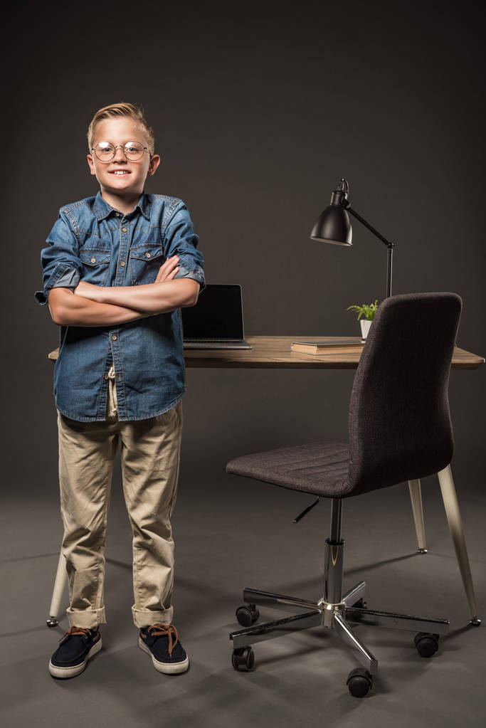 petit garçon en lunettes debout avec les mains croisées près de la table avec ordinateur portable, livre et lampe
  - Photo, image