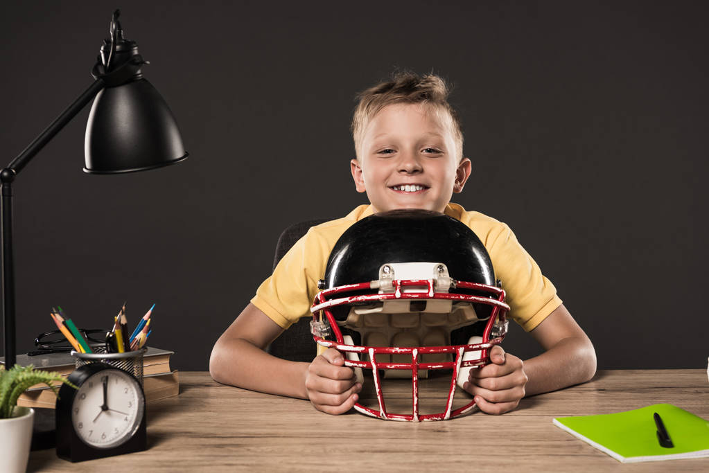 scolaretto felice con casco da calcio americano seduto a tavola con libri, piante, lampade, matite a colori e libro di testo su sfondo grigio
  - Foto, immagini