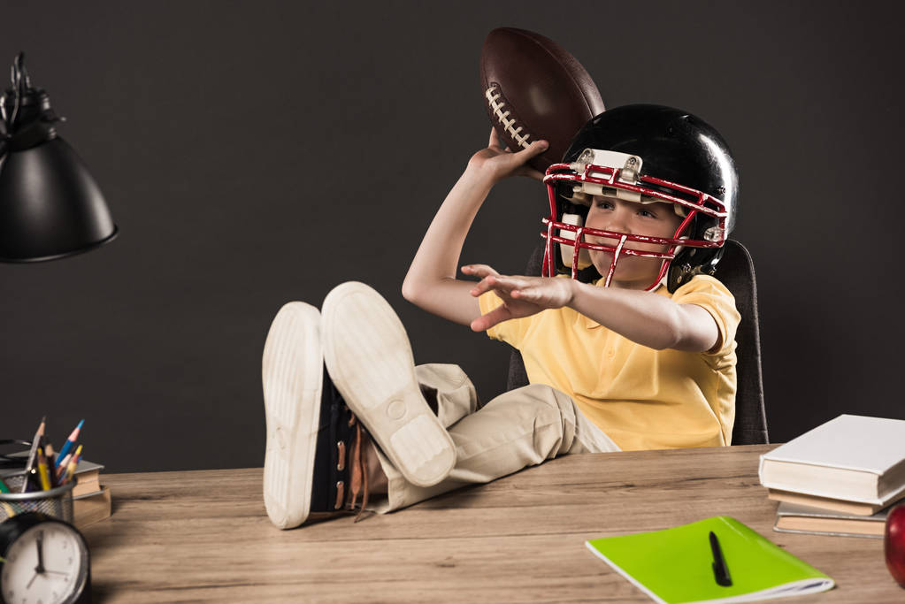 Schuljunge mit American-Football-Helm, der Ball wirft und mit Beinen auf einem Tisch mit Büchern, Pflanzen, Lampen, Farbstiften, Apfel, Uhr und Lehrbuch auf grauem Hintergrund sitzt  - Foto, Bild