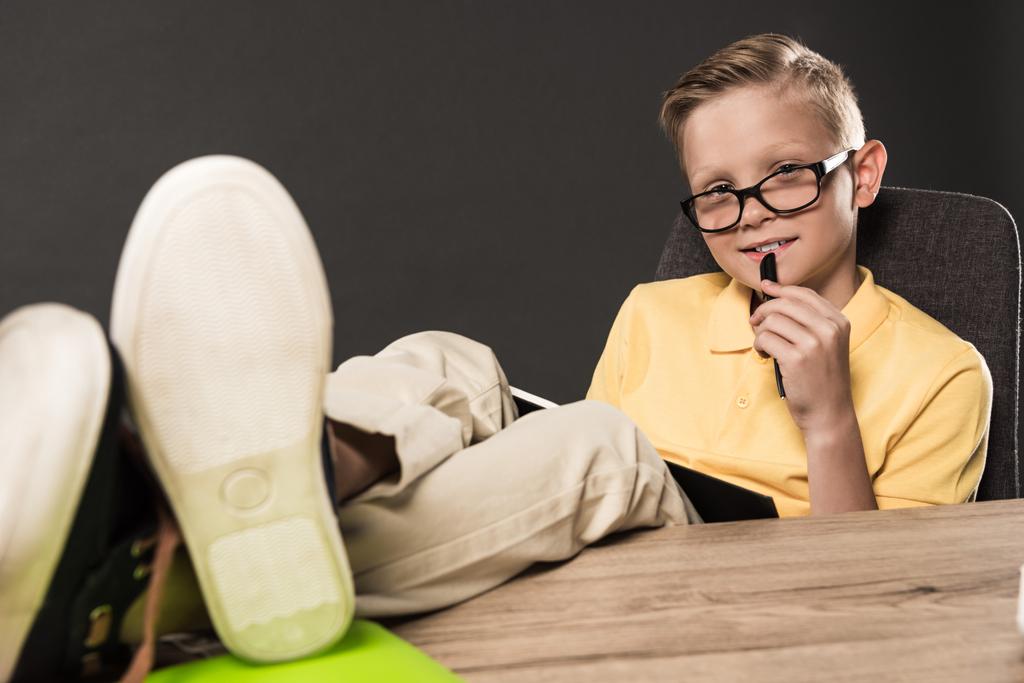 schoolboy in eyeglasses with textbook doing homework with legs on table on grey background  - Photo, Image