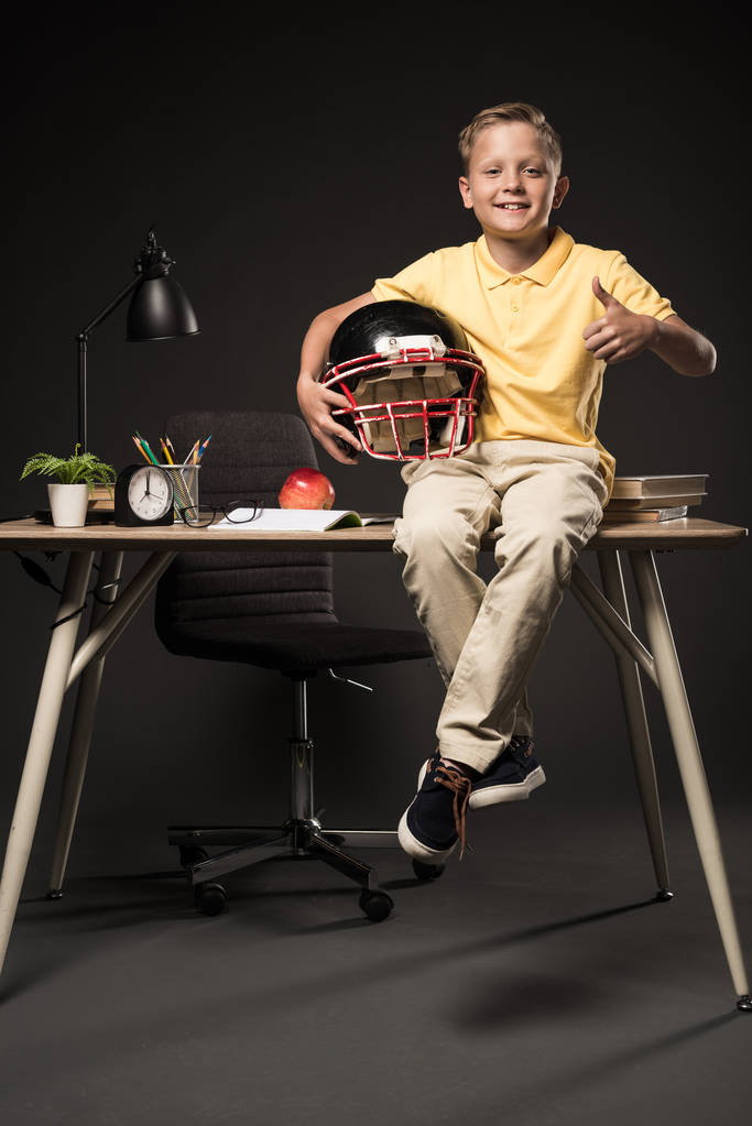 adorabile scolaro che tiene il casco da football americano, fa un gesto da pollice in su e siede sul tavolo con libri, piante, occhiali, lampada, matite di colore, mela, orologio e libro di testo su sfondo grigio
  - Foto, immagini