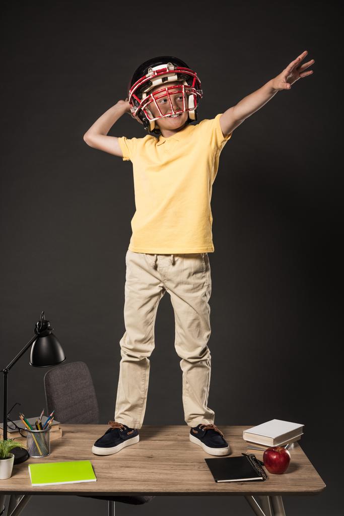 écolier souriant dans un casque de football américain imaginant qu'il lance le ballon et debout sur la table avec des livres, plante, lampe, crayons de couleur, pomme, horloge et manuel sur fond gris
  - Photo, image