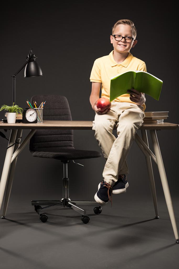 colegial sonriente sosteniendo manzana y haciendo los deberes mientras está sentado en la mesa con libros, plantas, lámparas, lápices de color, reloj y libro de texto sobre fondo gris
  - Foto, imagen