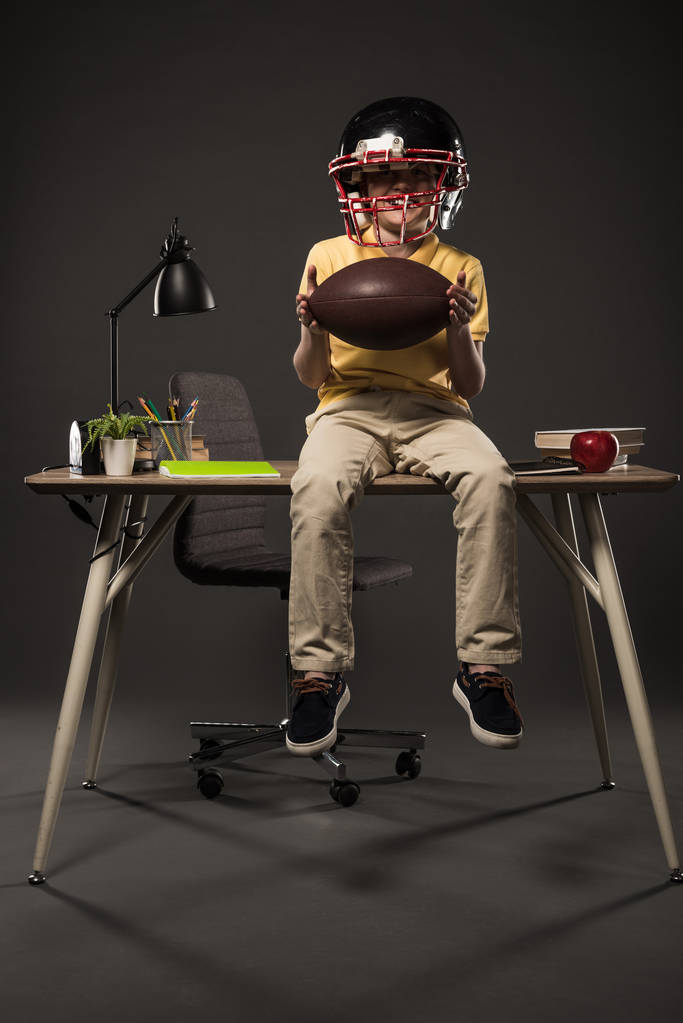scolaretto sorridente in casco da calcio americano che tiene la palla e siede sul tavolo con libri, piante, lampade, matite di colore, mela, orologio e libro di testo su sfondo grigio
  - Foto, immagini