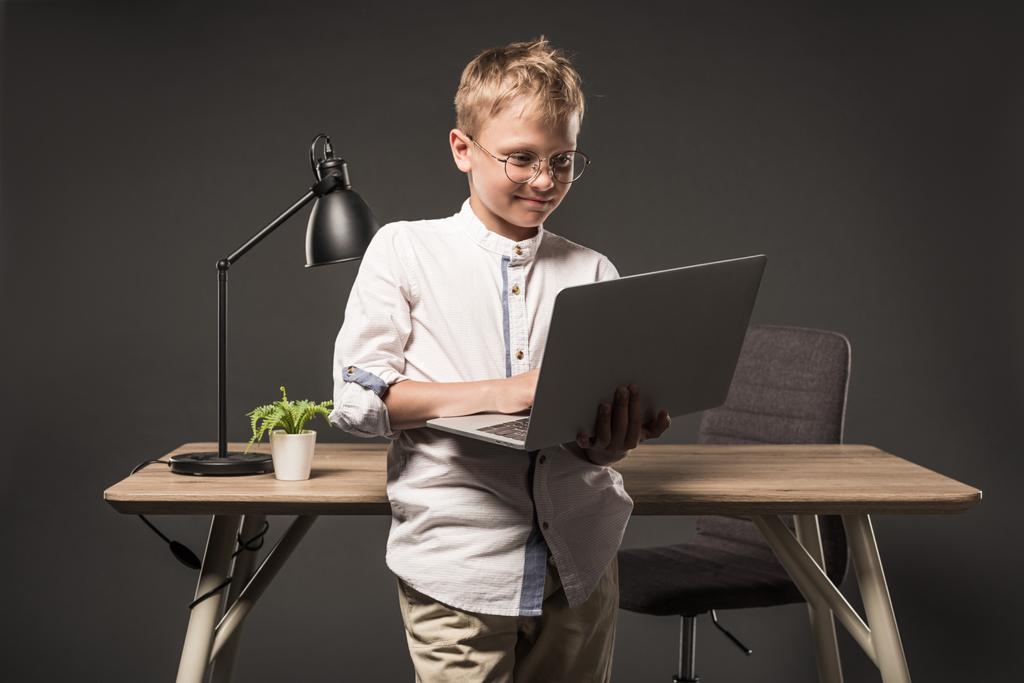 μικρό αγόρι σε γυαλιά που χρησιμοποιούν φορητό υπολογιστή κοντά στο τραπέζι με φυτό και φωτιστικό σε γκρίζο φόντο   - Φωτογραφία, εικόνα