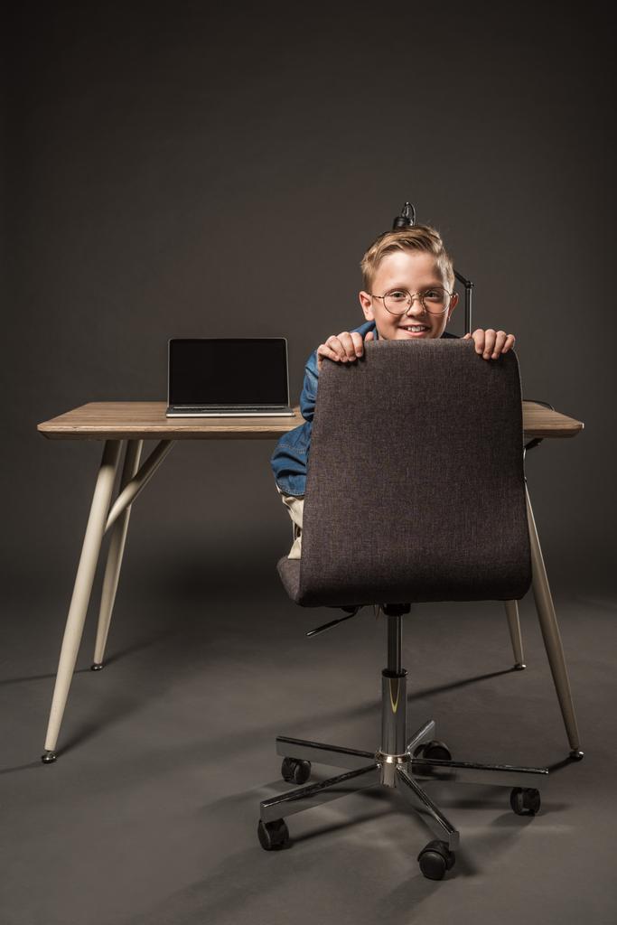 niño sonriente en anteojos sentado en la silla cerca de la mesa con el ordenador portátil y la lámpara sobre fondo gris
  - Foto, imagen