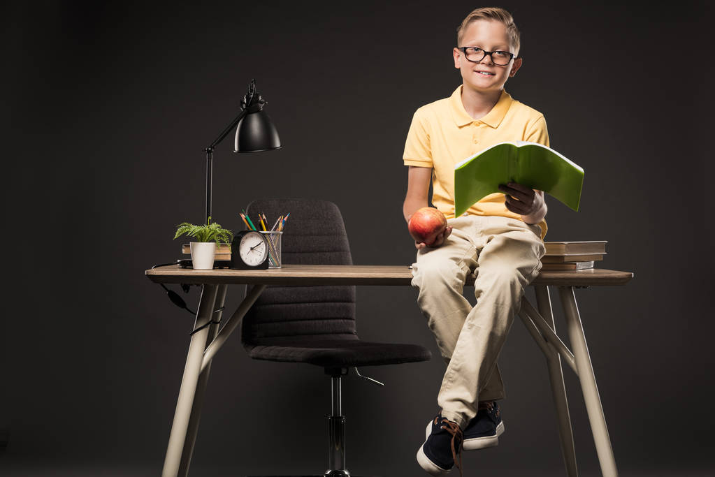 pieni koulupoika silmälaseissa pitämässä omenaa ja tekemässä läksyjä istuessaan pöydällä kirjojen, kasvien, lampun, värikynien, kellon ja oppikirjan kanssa harmaalla taustalla
  - Valokuva, kuva