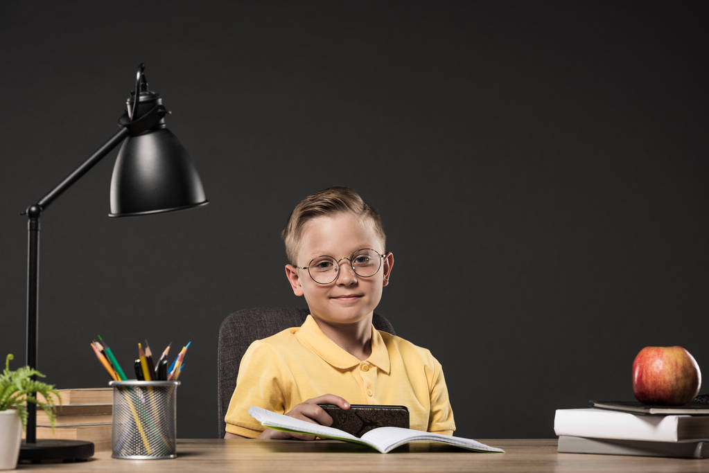 Lächelnder Schuljunge mit Smartphone am Tisch mit Büchern, Pflanze, Lampe, Farbstiften, Apfel, Uhr und Lehrbuch auf grauem Hintergrund  - Foto, Bild