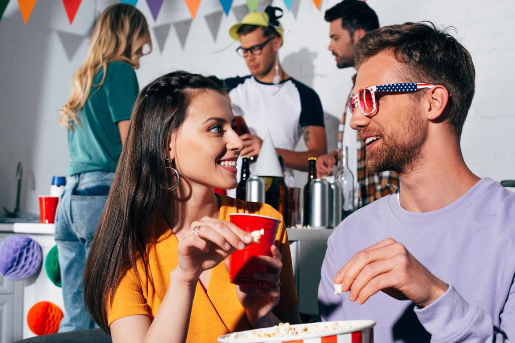 šťastný mladý muž a žena vzájemně usmíval, zatímco pít pivo a jíst popcorn na domácí party - Fotografie, Obrázek