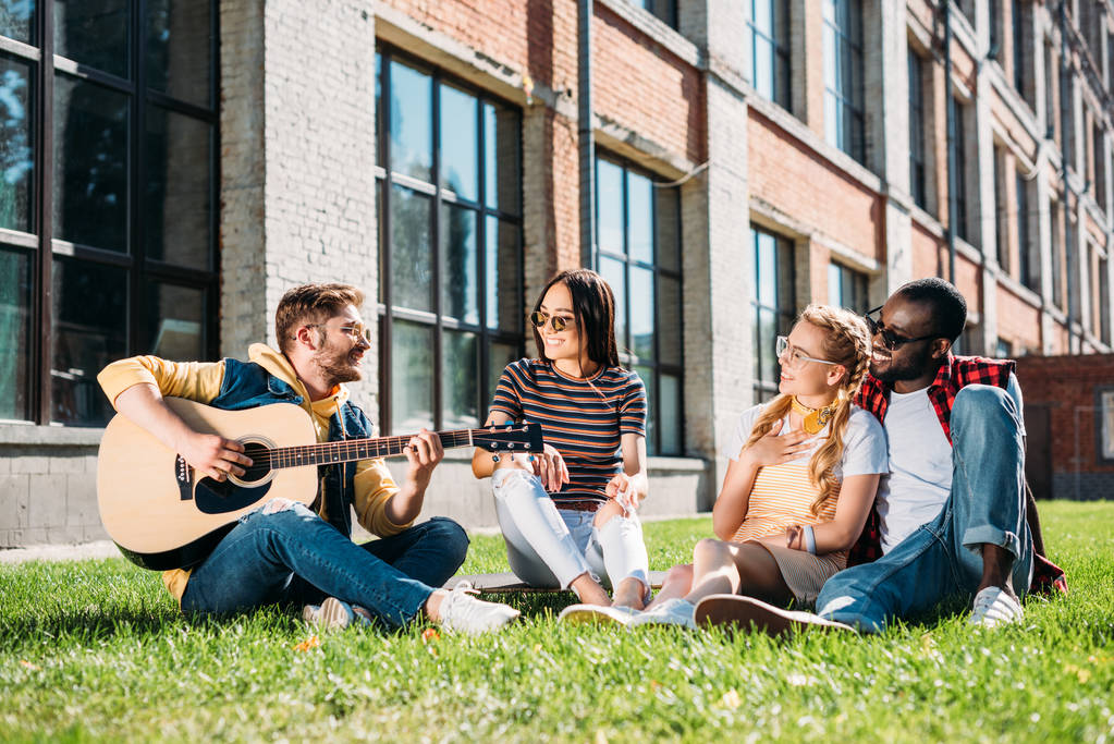 夏の日の緑の芝生で休んでアコースティック ギターと友達の異人種間のグループ - 写真・画像