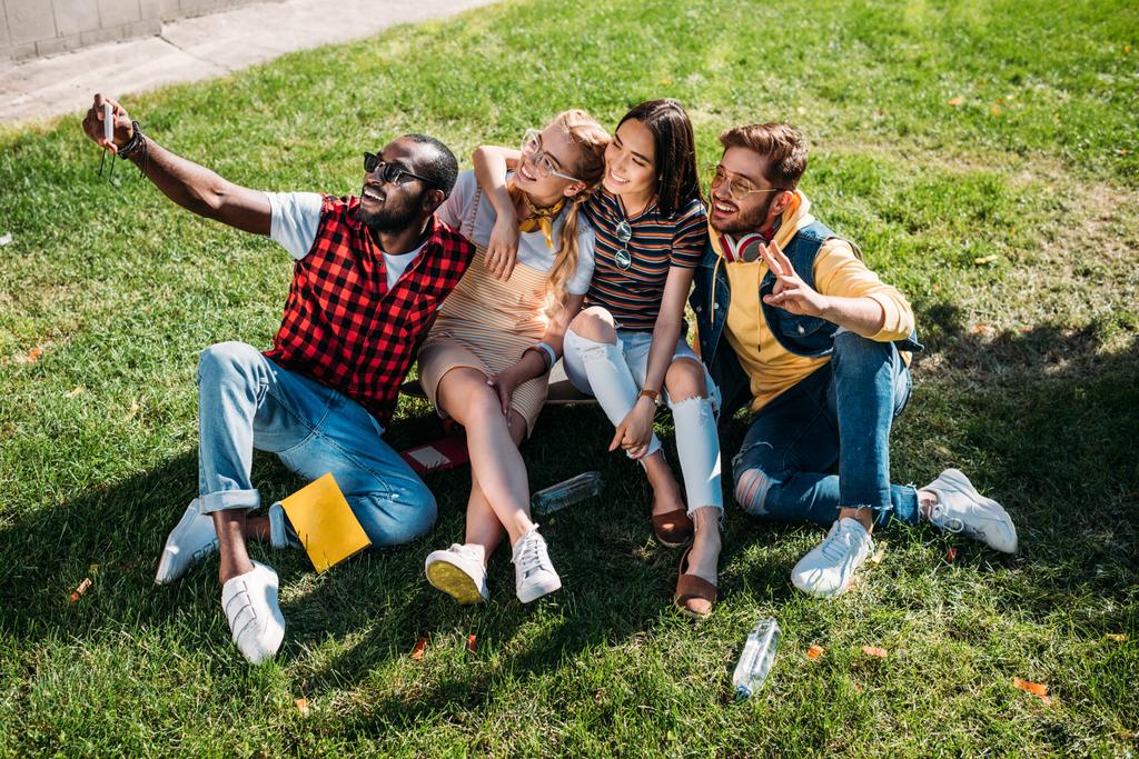 мультиэтнические друзья делают селфи на смартфоне, отдыхая на зеленой лужайке вместе
 - Фото, изображение