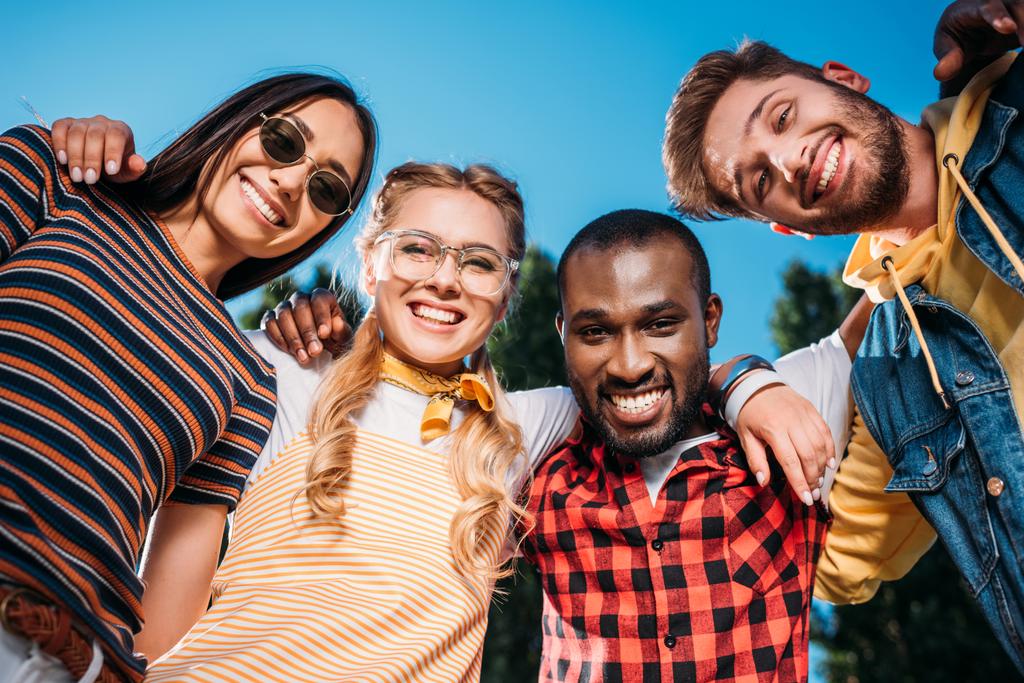 Onderaanzicht van glimlachen multiraciale jonge vrienden op zoek naar camera met blauwe hemel op achtergrond - Foto, afbeelding