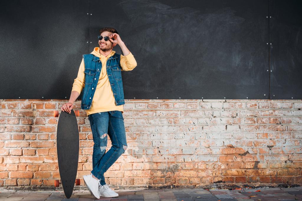 homme élégant dans des lunettes de soleil avec longboard debout sur la rue
 - Photo, image