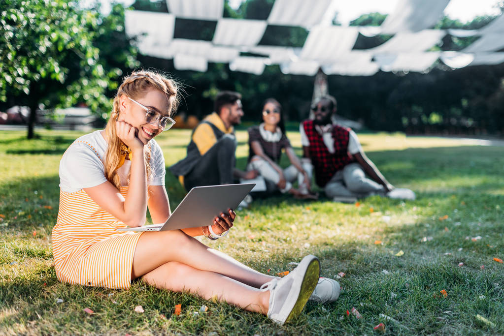 selective focus of young smiling woman with laptop and multiracial friends behind resting on green grass in park - Photo, Image