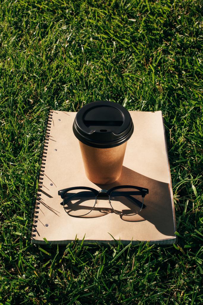 close up view of coffee to go, notebook and eyeglasses on green grass - Photo, Image