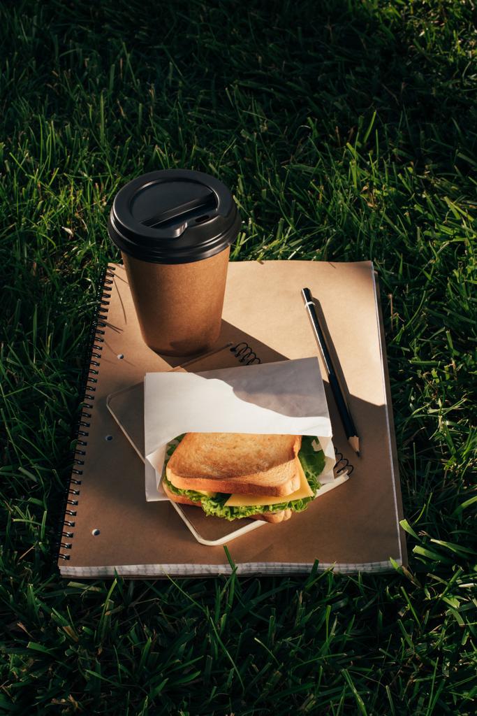 κλείνω πάνω θέα σημειωματάρια, σάντουιτς και καφέ για να πάει στο πράσινο χορτάρι - Φωτογραφία, εικόνα