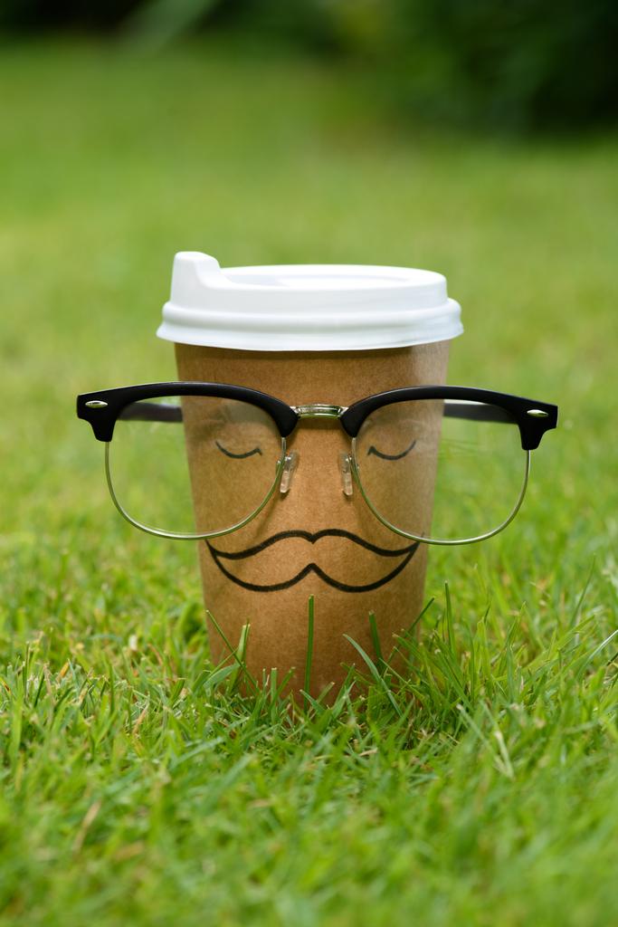 zbliżenie jednorazowego kubka z znak wąsy i okulary na zielony trawnik - Zdjęcie, obraz