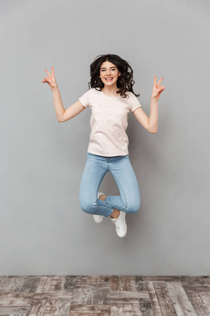 Photo de jolie jeune femme mignonne sautant isolée par-dessus un mur de fond gris montrant un geste de paix
. - Photo, image