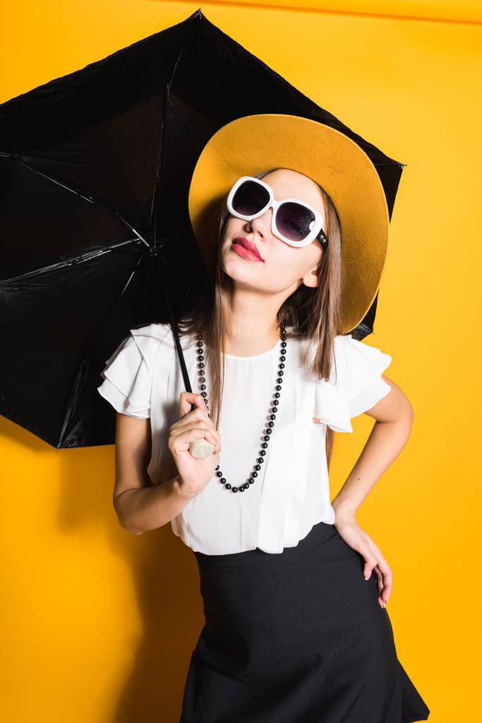 atrakcyjny, stylowa dziewczyna w okulary przeciwsłoneczne i kapelusz posiada parasol i pozowanie na żółtym tle - Zdjęcie, obraz