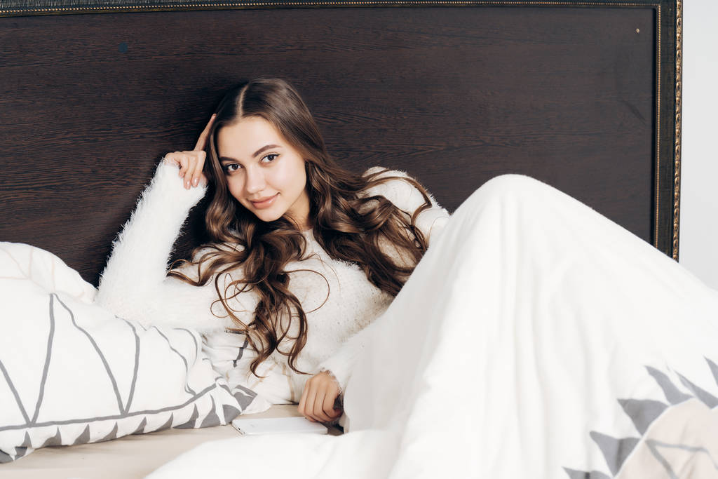 мила довгошерста дівчина в білій піжамі лежить на ліжку, відпочиваючи після робочого дня і посміхаючись
 - Фото, зображення