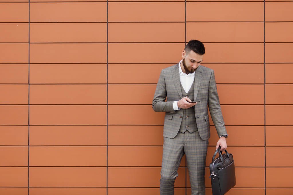 бизнесмен в костюме и с помощью современного смартфона возле офиса рано утром, успешный работодатель, чтобы заключить сделку, стоя рядом с небоскребом
 - Фото, изображение
