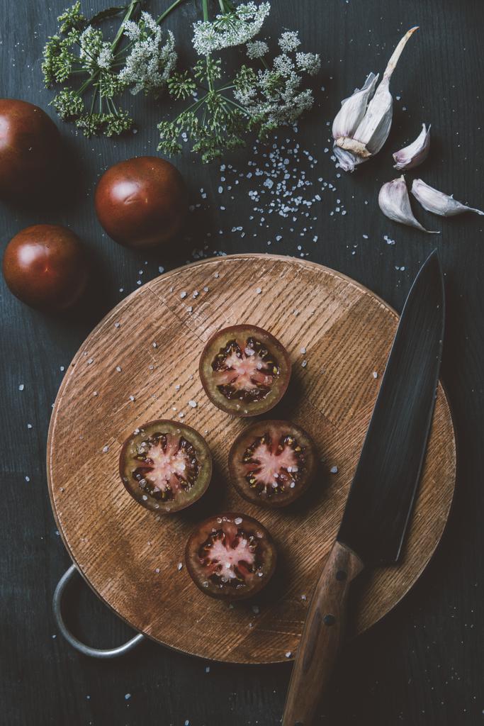 vista superior de los tomates príncipe negro en la tabla de cortar con cuchillo, ajo, sal y flores
 - Foto, imagen