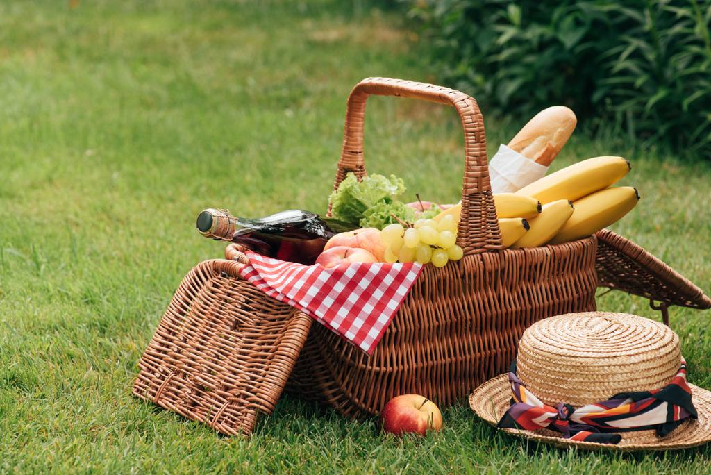 yummy fruits and bottle of champagne in wicker basket on green grass at picnic - Photo, Image