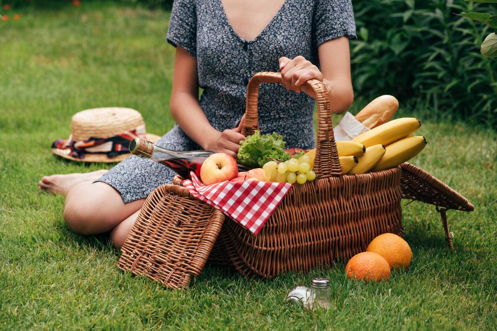 skadrowana kobieta siedzi na zielonej trawie, piknik i dotykając wiklinowy koszyk - Zdjęcie, obraz