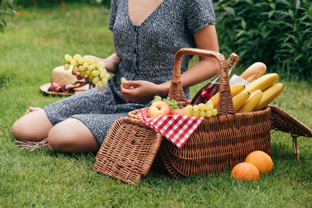 cropped image of woman eating grapes and sitting on green grass at picnic - Photo, Image