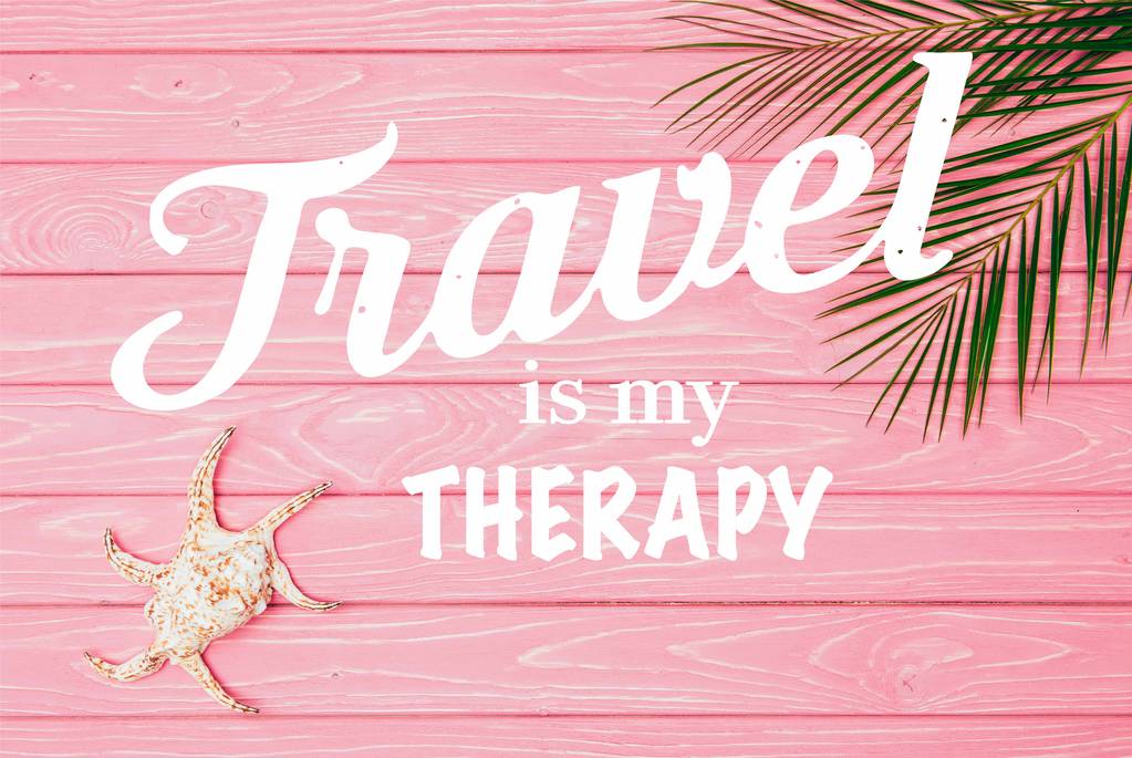 Draufsicht auf Muschel mit Palmblättern auf rosa Holzoberfläche mit dem Schriftzug "Reisen ist meine Therapie"  - Foto, Bild