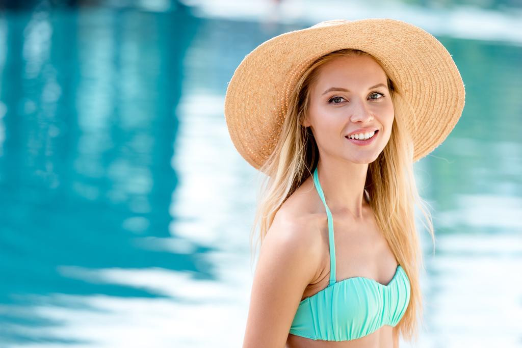 приваблива молода жінка в солом'яному капелюсі і бікіні дивиться на камеру біля басейну
 - Фото, зображення