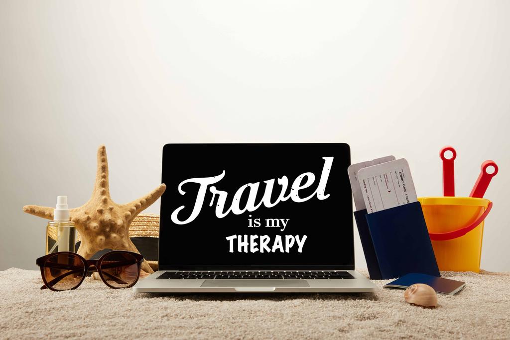 close-up vista de laptop com "viajar é a minha terapia" lettering, estrela do mar, óculos de sol, passaportes com bilhetes e balde de brinquedo na areia em fundo cinza
 - Foto, Imagem