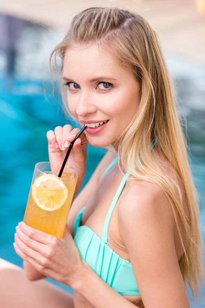 hermosa joven con refrescante bebida naranja mirando a la cámara junto a la piscina
 - Foto, Imagen