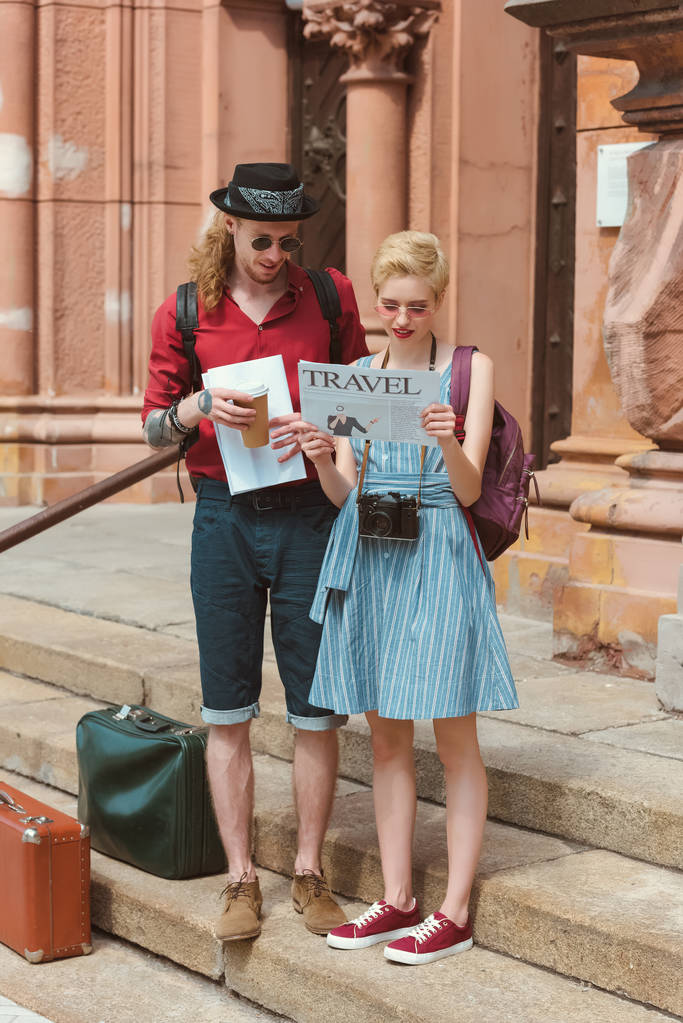 un par de turistas con café leyendo el periódico de viaje mientras están de pie en las escaleras con maletas retro
 - Foto, Imagen