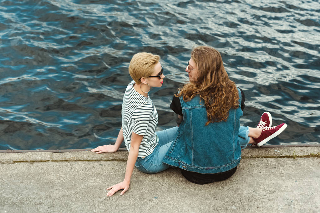 Hochwinkelaufnahme von Freund mit Tätowierungen und stilvoller Freundin, die auf Brücke sitzt und einander ansieht - Foto, Bild