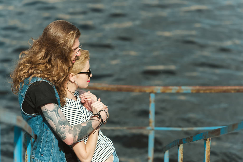 вид збоку на хлопця з татуюваннями, що обіймає стильну дівчину на мосту біля річки
 - Фото, зображення