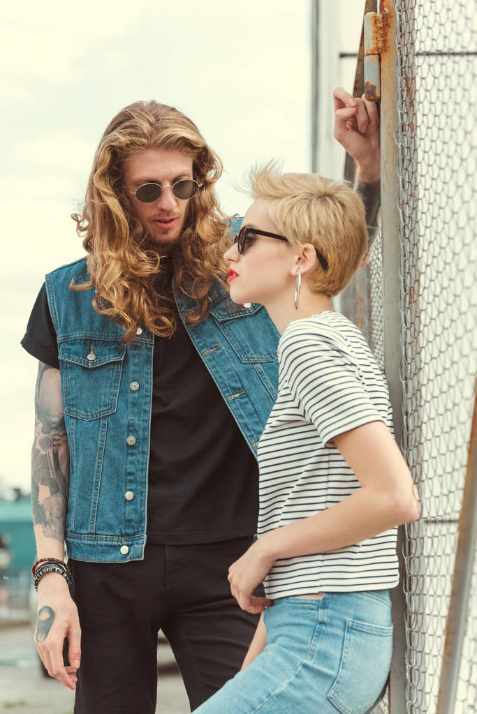 boyfriend with tattoos leaning on fence and looking at stylish girlfriend - Photo, Image