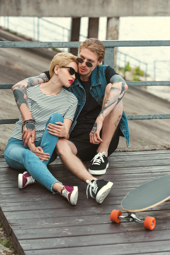 poikaystävä tatuoinnit ja tyylikäs tyttöystävä istuu sillalla ja halaus lähellä rullalauta
 - Valokuva, kuva