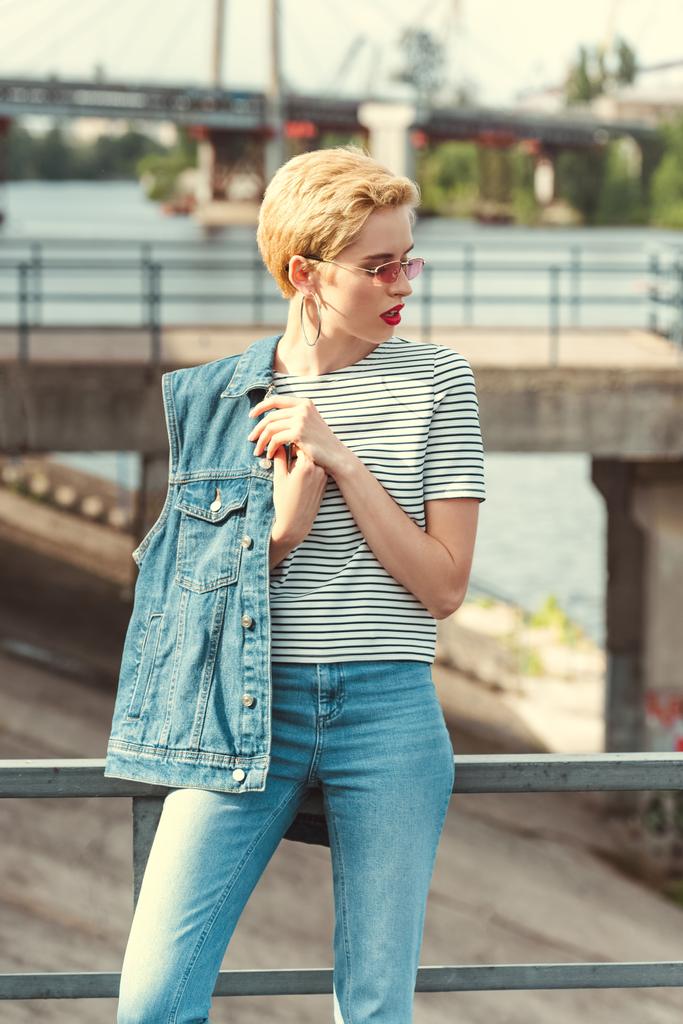 stylish girl standing on bridge in jeans and denim vest and looking away - Photo, Image