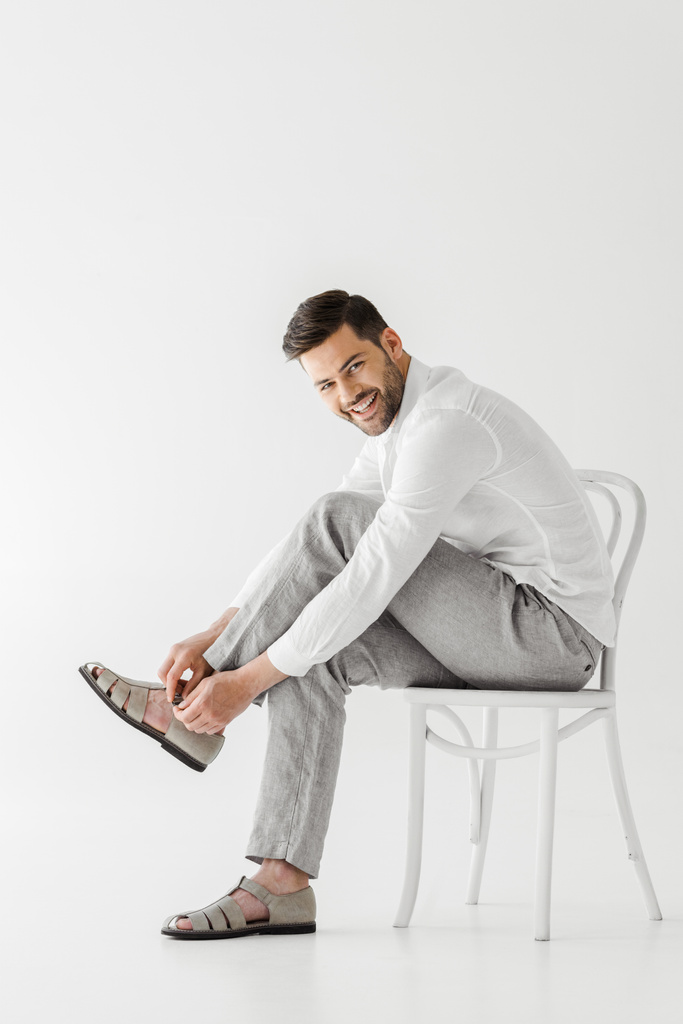 улыбающийся мужчина в льняной одежде сидит на стуле и надевает сандалии изолированные на сером фоне
  - Фото, изображение